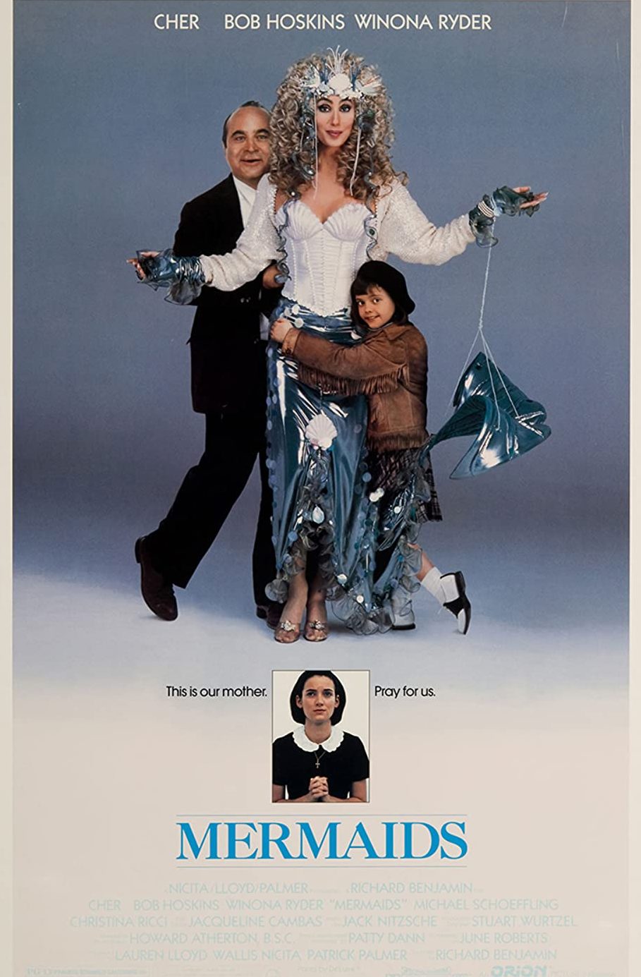a blue grey background that fades to cream in the lower third. A woman in a mermaid costume smiling to the camera. She is being hugged by a small girl in a brown fringed jacked and a man in a suit. Under their feet is a photo of a girl praying in a black dress. Text: This is our mother. Pray for us. Mermaids