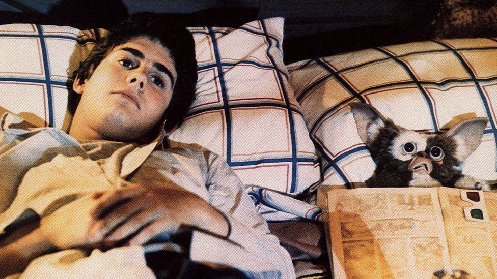 billy peltzer and gizmo lie in bed watching a film. billy is a teenage boy and looks bored. gizmo is a mogwai and looks scared. he holds a comic and 3d glasses.